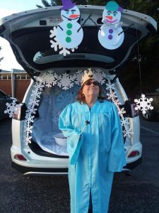 Melody Turner in a blue choir robe, holding a baton with a start on it. The car behind her is decorated with cut out snowflakes and two poster board snowmen.