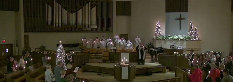 Christmas Eve Candlelight Service in the worship center. The room is lit by small candles held by attendees with as white lights glow on Christmas trees.