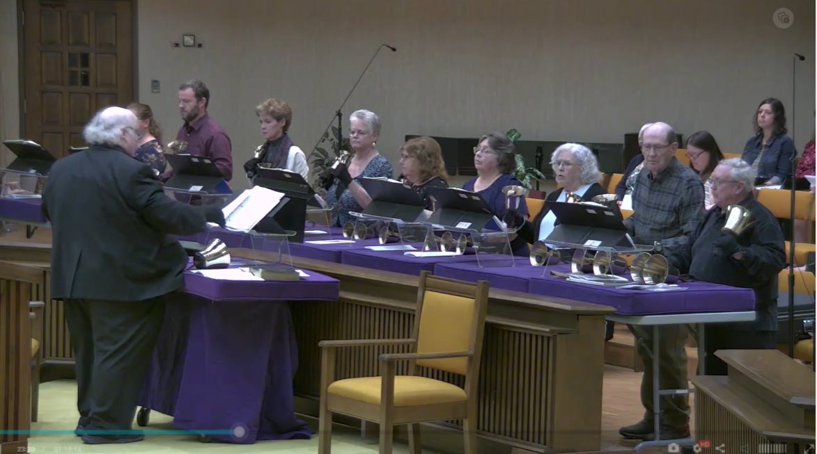 Tim Bounds directs nine members of a handbell choir. Tables with purple padding hold the bells and music folders.