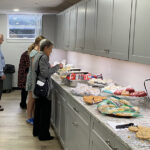 Three women select food from a kitchen counter, while a man with white hair waits in line.